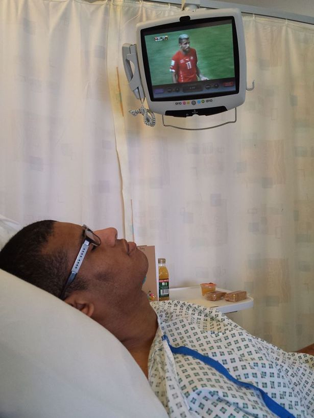 Mixed race man wearing glasses lies in a hospital bed with TV on in the background. His eyes are closed.