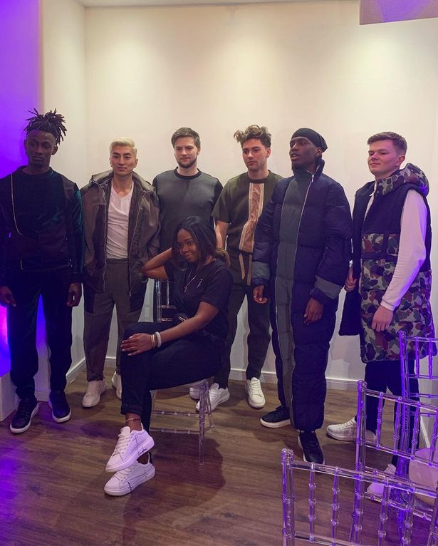 Six male models wearing dark clothes stand around Cherise who is sitting cross leg in a chair.