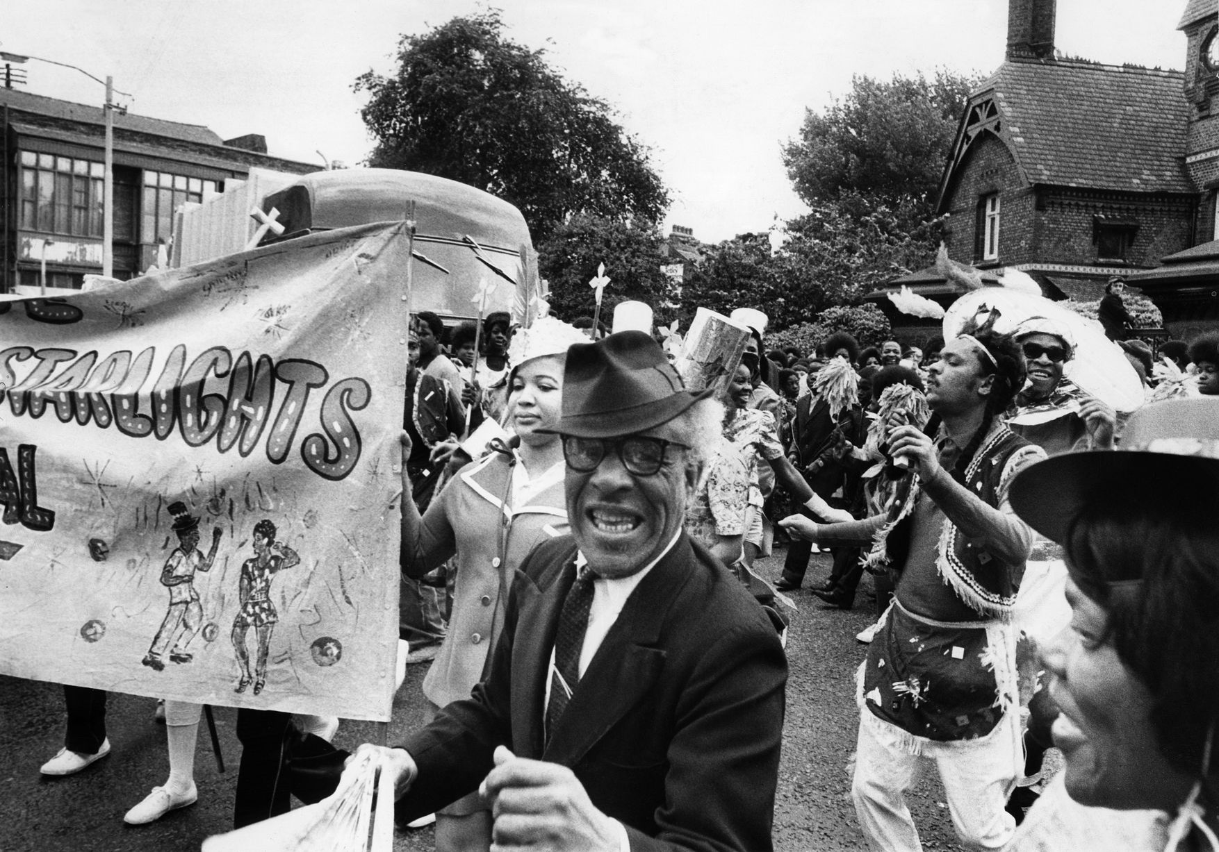 Black and white photo of the carnival procession. Man smiling wearing a hat. Steel bands and brightly dressed Indian dancers lead the way for the traditional lorry borne floats in the Moss Side procession
