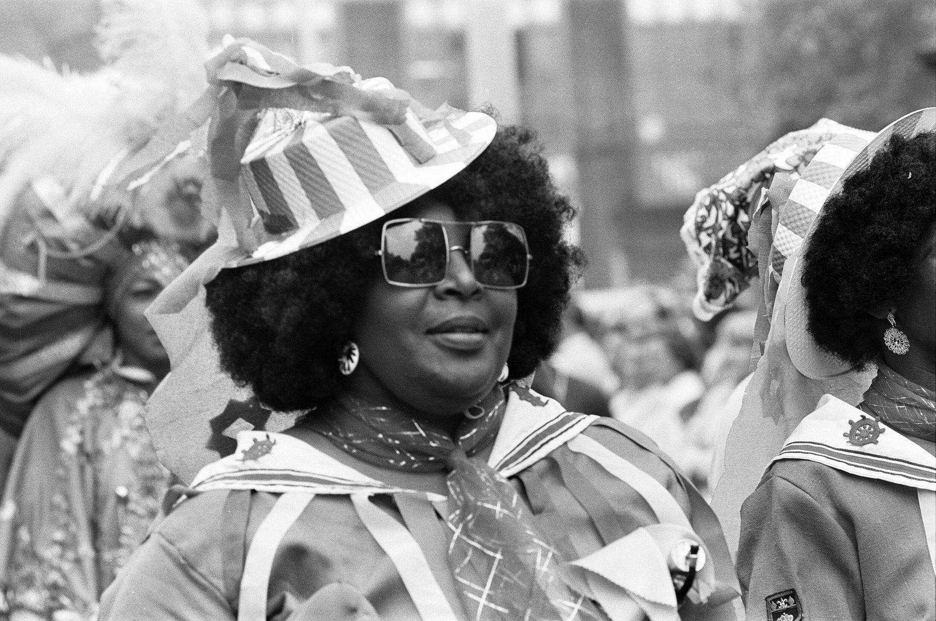 Lady with afro hair and a fancy hat and sunglasses smiles during the carnival.