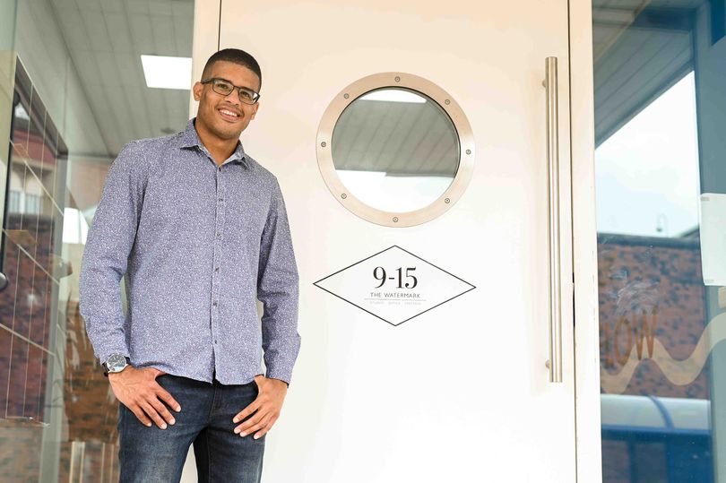Tall mixed race man with glasses stands in front of a white door with his hands in his pockets. He has a big smile on his face.